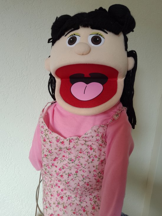 Handpop met open mond, bruine ogen, zwart haar in knotjes. Draagt roze trui en bloemenjurk.