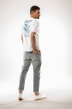 Man met op de rug een witte T-shirt met blauw blad-print "CHIEF". Grijze jeans.