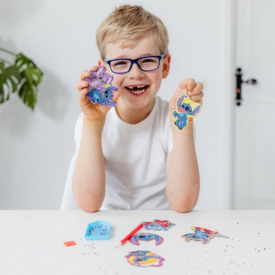 Un garçon avec des lunettes tient deux magnets Stitch diamond painting terminés : un avec deux Stitch violets et un avec Stitch aux accents jaunes.