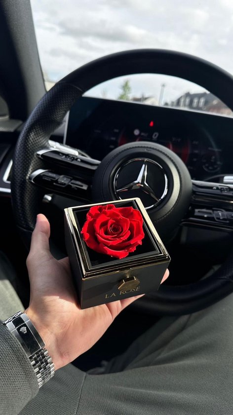 Een hand met een horloge houdt een zwarte juwelendoos vast met een gouden rand en een rode roos erin. Een auto stuur is zichtbaar op de achtergrond.