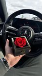 Een hand met een horloge houdt een zwarte juwelendoos vast met een gouden rand en een rode roos erin. Een auto stuur is zichtbaar op de achtergrond.