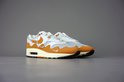 Paar witte met oranje Nike Air Max 1 Patta sneakers met beige en bruine accenten, op een grijze achtergrond.