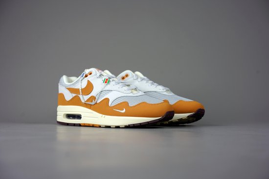 Paar witte met oranje Nike Air Max 1 Patta sneakers met beige en bruine accenten, op een grijze achtergrond.