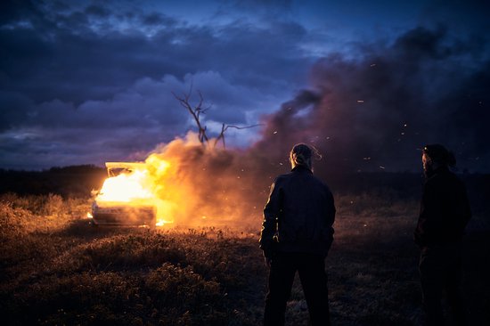 Twee mannen kijken naar een brandende auto in een veld bij schemering. Rook en vonken stijgen op.