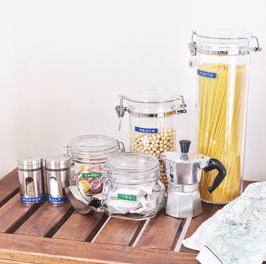 Glazen potten met etenswaren op een houten plank. Gelabeld met 'PASTA', 'BEANS', 'CANDY', 'TEA', 'SALT', 'PEPPER'. Ook een koffiezetapparaat.