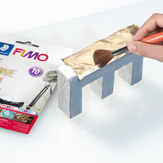 Een hand brengt gouden bladmetaal aan op een grijs en wit E-vormig object met een kwast.
