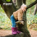 Meisje met blonde staart, teddy jas en paarse laarzen, leunend op een boom in een bos.