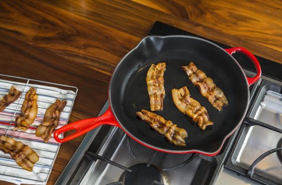 Kersrode gietijzeren koekenpan met vijf plakjes krokant gebakken spek erin en op een rooster ernaast.