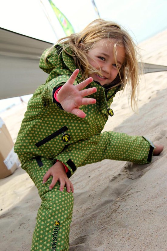 Meisje met blonde haren, zittend op het zand, met een groene jas en broek met sterrenprint.