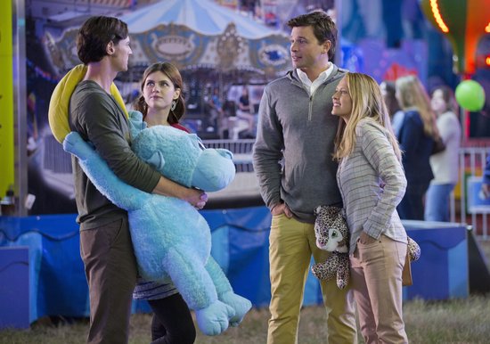 Twee koppels staan op een kermis. Een man houdt een grote blauwe knuffelbeer vast. De ander houdt een kleine bruine luipaard knuffel.