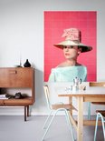 Audrey Hepburn portant un grand chapeau orné d'un nœud rose, un haut pâle, sur fond rose, dans un salon avec buffet en bois et tables.