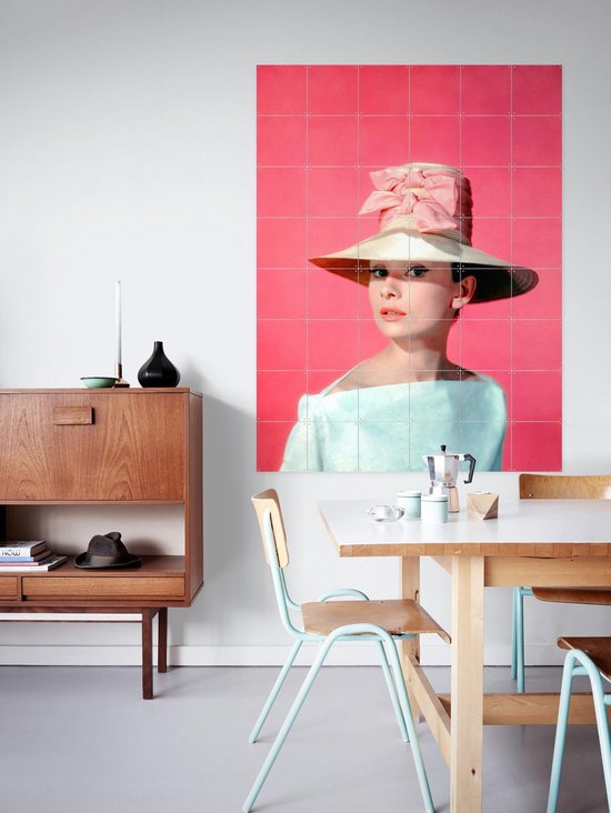 Audrey Hepburn portant un grand chapeau orné d'un nœud rose, un haut pâle, sur fond rose, dans un salon avec buffet en bois et tables.