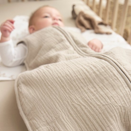 Zicht op een baby in een zandkleurige, gevoerde slaapzak. De slaapzak heeft een gebreide structuur.