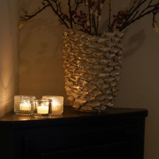 Twee brandende C-lights theelichtjes in glas met reliëf, naast een grote vaas met decoratieve takken.