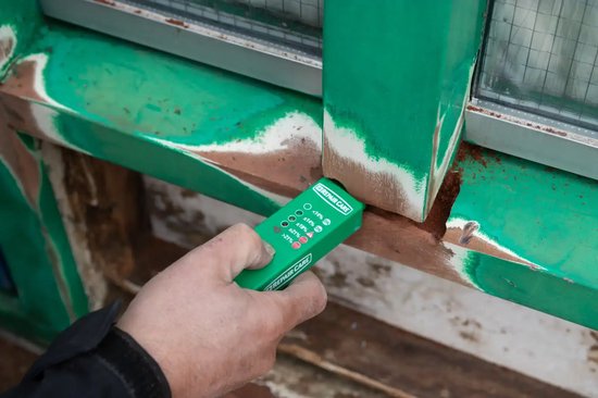 Groene Easy.Q vochtmeter met deksel in de hand, naast groen geschilderd hout met bruine plekken.