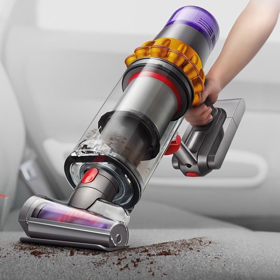 Een hand houdt een paarse, gele en zilveren steelstofzuiger vast, die vuil opzuigt van een stoffen autostoel.