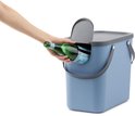 Blauwe afvalbak met grijze deksel. Hand wordt gebruikt om een fles in de opening te doen.