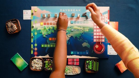 Twee personen spelen het bordspel Daybreak. Het spelbord toont een wereldkaart met pionnen. Rondom het bord liggen schaaltjes met speelstukken.