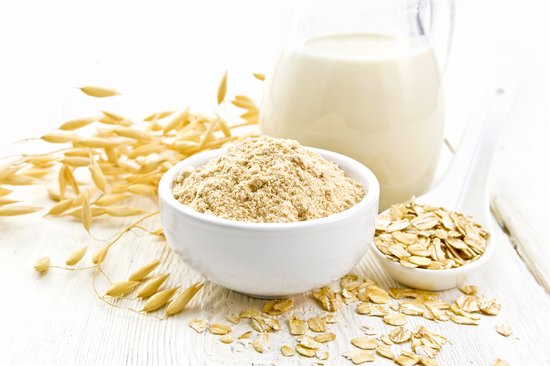 Witte kom met havermoutpoeder, oren van haver, een lepel met havermoutvlokken en een karaf melk.