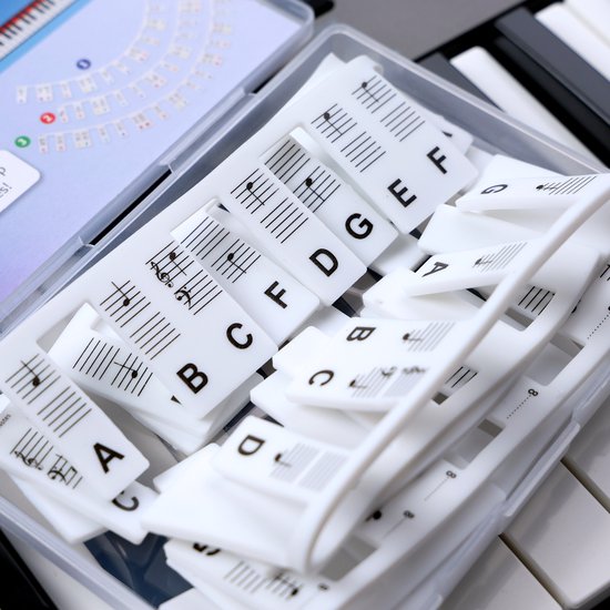 Witte pianotoetsen met daarop vele witte stickers met muzieknoten en letters zoals A, B, C, D, E, F, G. De stickers liggen in een transparant doosje.