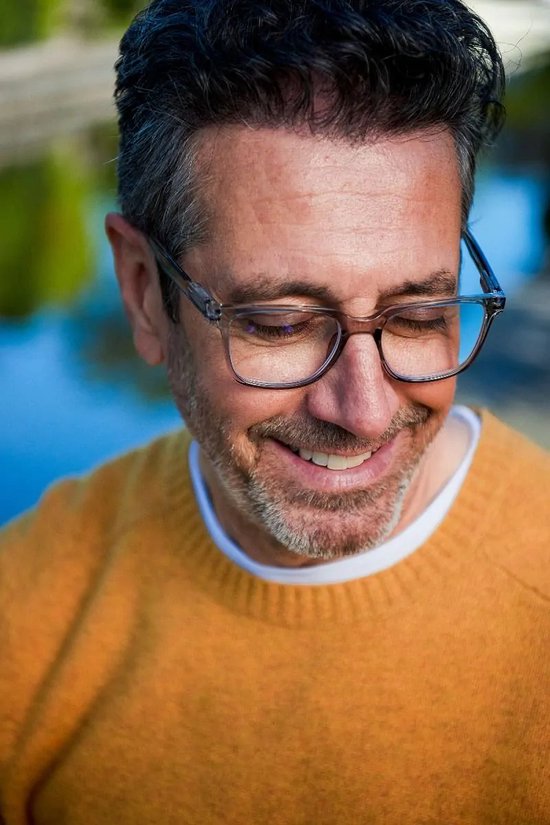 Homme portant des lunettes de lecture à monture transparente, un pull tricoté jaune, regardant un téléphone.