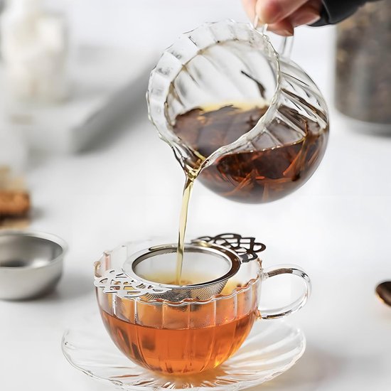 Water wordt gegoten uit een geribbelde glazen theepot door een zilverkleurige theezeef met vleugel handvatten in een glazen kop.