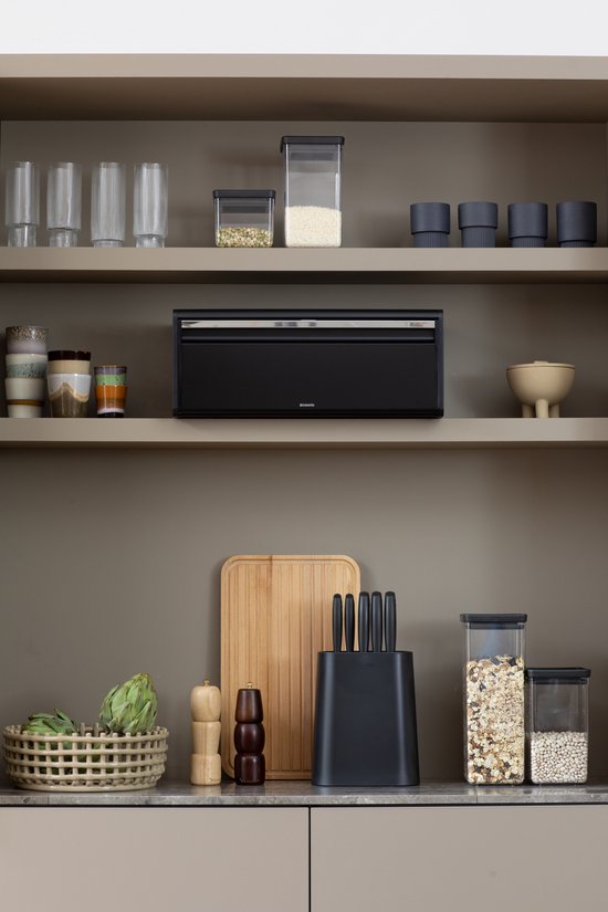 Boîte à pain Brabantia noire avec couvercle à rabat, posée sur un comptoir entre des accessoires de cuisine.