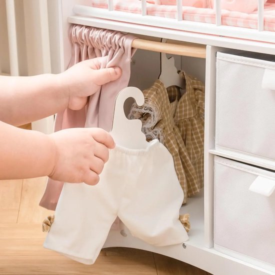 Kindhanden hangen witte poppenbroek aan een houten hanger in het poppenbedje, naast poppenkleding in de opbergvakken.