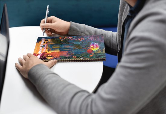 Een persoon met een pen schrijft in een 'Magische Tuinen Kleurboek' van Colourific Moments op een tafel.