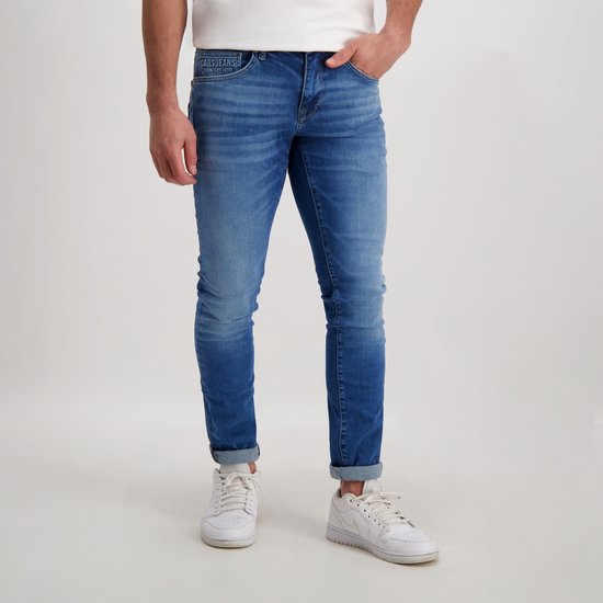 Man in een medium blauwe spijkerbroek met opgerolde zoom en witte sneakers. De broek heeft het Cars Jeans logo op de tailleband.