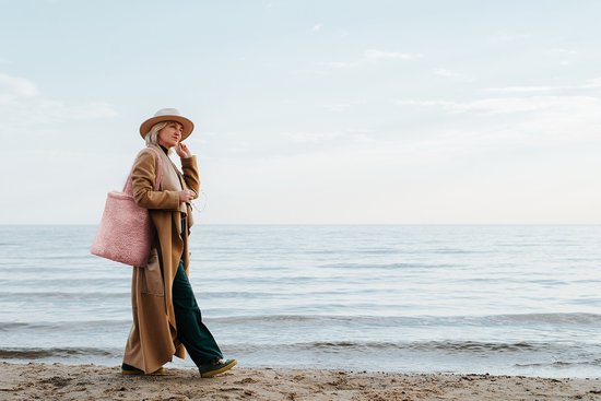 Femme marchant sur la plage avec un sac teddy rose et un manteau beige.
