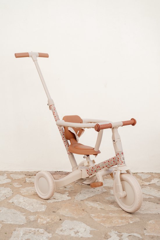 Crèmekleurige 4-in-1 driewieler met duwstang, bruine zitting en handvatten, op stenen ondergrond. Frame met bloemenprint.