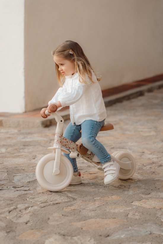 Meisje met blonde staartjes in witte blouse en spijkerbroek zit op een crèmekleurige driewieler met bloemenprint.