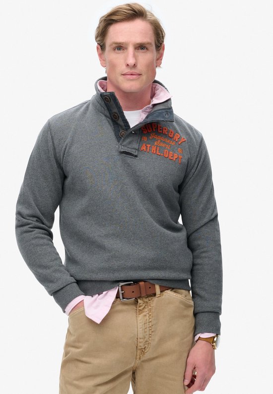 Man met blond haar, staand met hand in de zak, in een grijze Superdry sweater met oranje opdruk, roze hemd en beige broek.
