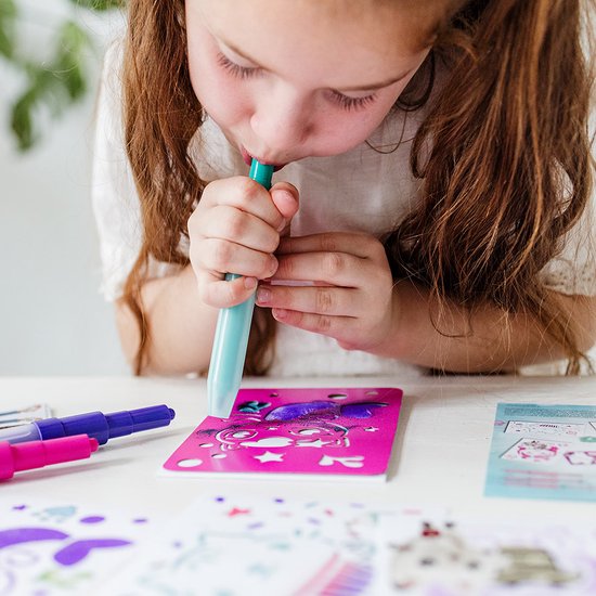 Meisje met bruine staartjes gebruikt een lichtblauwe spray pen op een roze Gabby's Poppenhuis sjabloon met katten- en glitter-elementen.