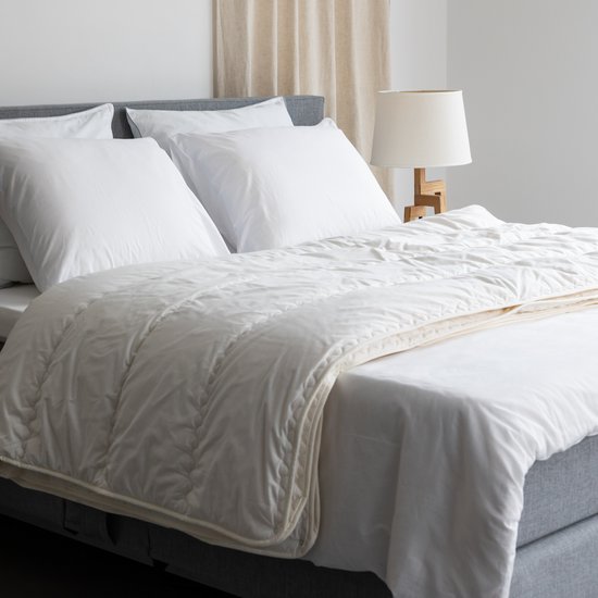 Wit, gewatteerd dekbed op een opgemaakt bed met witte lakens en kussens. De vulling is zichtbaar door de naden.