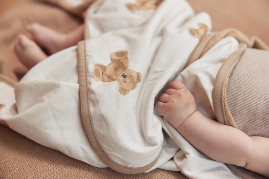 Pieds et main de bébé sous une cape de bain blanc cassé avec imprimé ours en peluche et bordure beige.