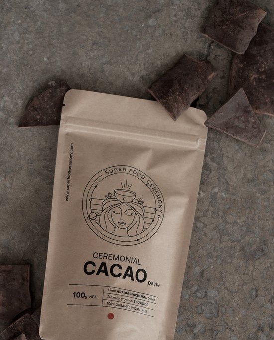 Bruine papieren zak met 'Ceremonial Cacao paste' logo, omringd door cacaostukken op een grijze ondergrond.