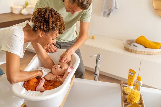 Een moeder en vader badderen hun baby in een ligbad. De baby draagt een oranje zwembroekje.