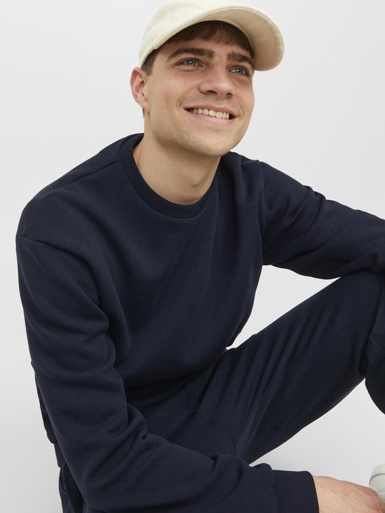 Model in een donkerblauwe sweater en joggingbroek, met een beige pet.