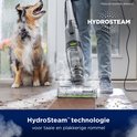 Een hond kijkt toe hoe de HydroSteam stofzuiger met stoom hardnekkig vuil op een houten vloer reinigt.