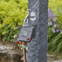 GARDENA besproeiingscomputer op stenen pilaar bij kraan in de regen. Tuin op achtergrond.