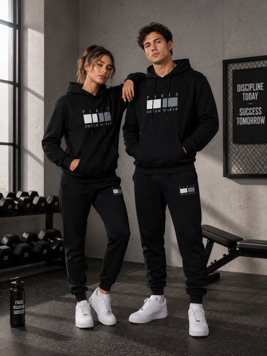 Een man en vrouw dragen zwarte "PARIS DREAM MAKER" trainingspakken in een fitnessruimte.