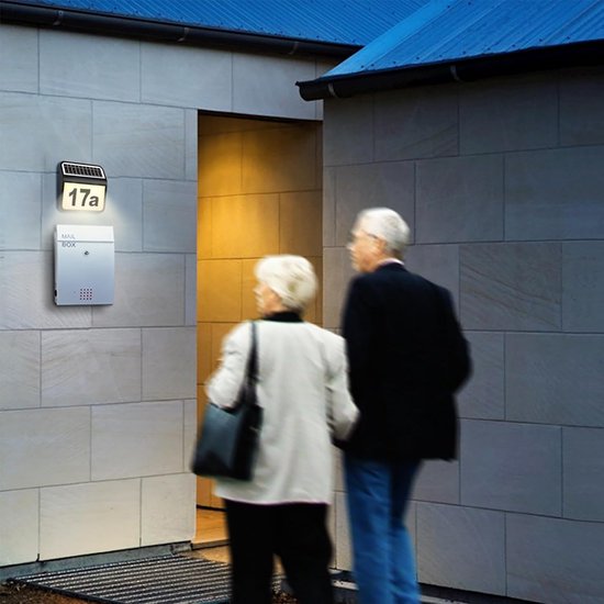Ouder stel loopt naar een verlicht huisnummerbord '17a' op een lichte stenen muur.