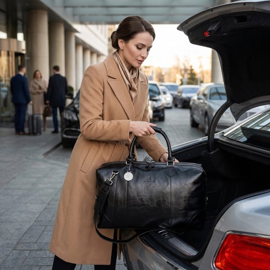Vrouw met een bruine jas en sjaal plaatst een zwarte leren reistas in de kofferbak van een auto.