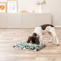 Een bruine puppy met witte vlekken zoekt naar lekkernijen in een grijze, witte en groene snuffelmat op een houten vloer.