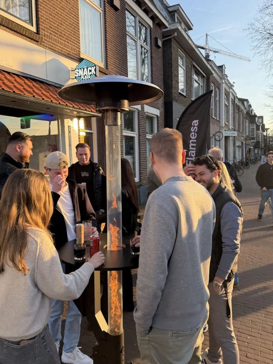 Groep mensen rond een brandende terrasverwarmer met tafel en 'SNACK PLAZA' uithangbord.
