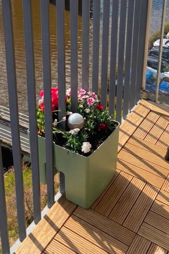 Lichtgroene plantenbak met rode en roze bloemen, hangend aan een grijze balustrade boven houten vloerplanken.