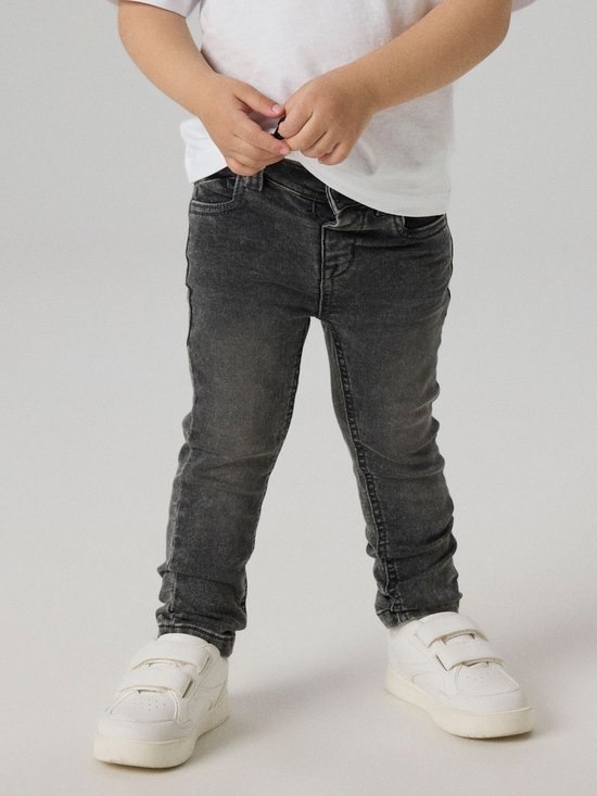 Onderlichaam van een jongen in een witte T-shirt en donkergrijze spijkerbroek.