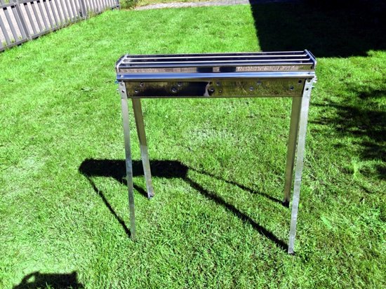 Een glimmende metalen sate grill met drie stangen, staand op groen gras met schaduw.
